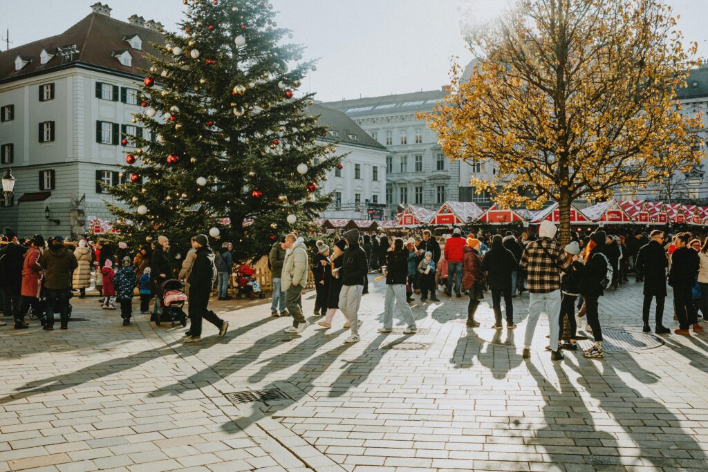 Crowds enjoy a festive Christmas market in a European city, with holiday decorations and a large Christmas tree.